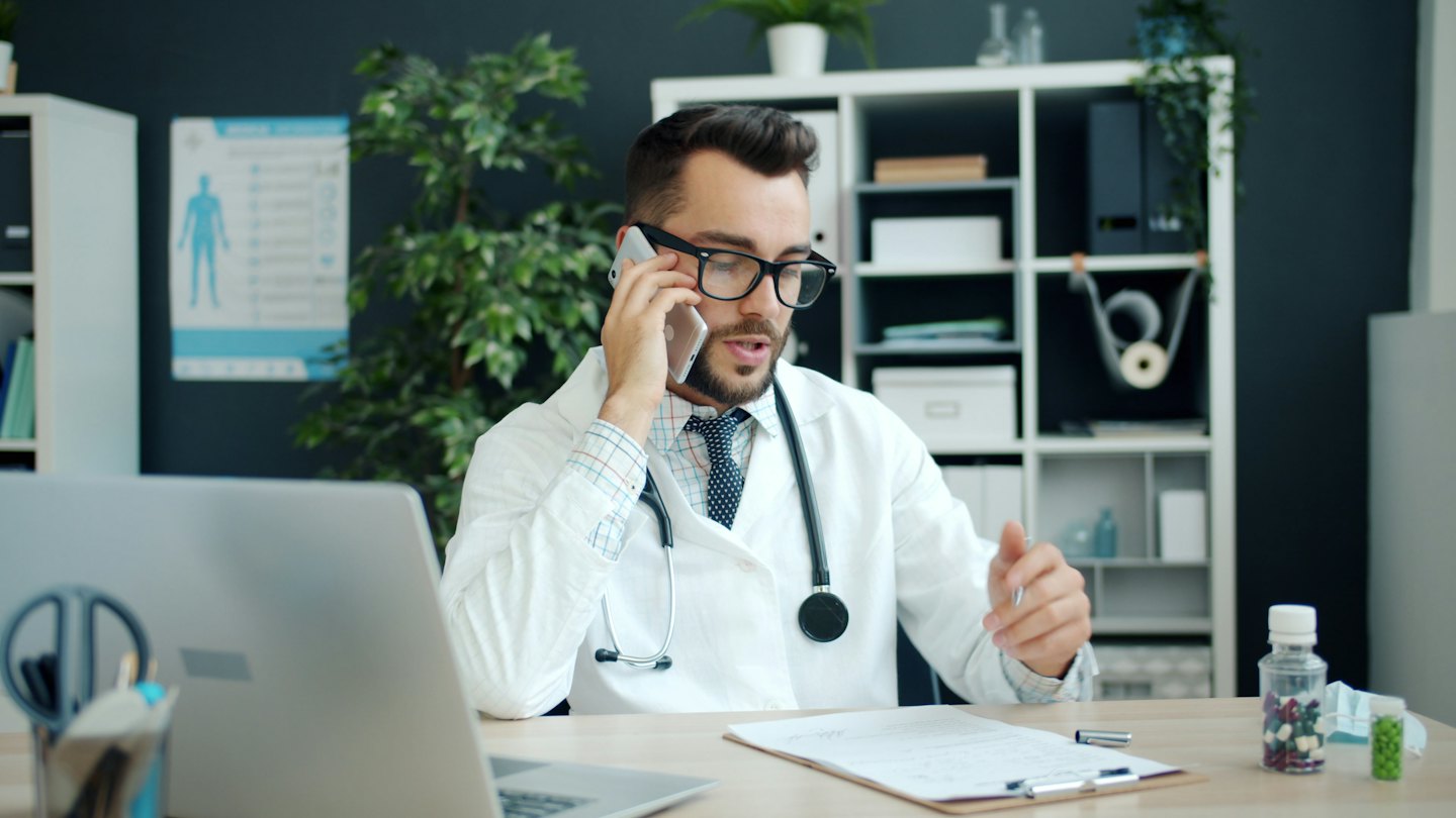 Medico al telefono alla sua scrivania mentre esamina la cartella di un paziente.
